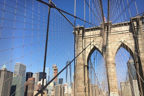 An intricate web of cables and suspension wires frames the stone arches of the Brooklyn Bridge, with the skyline of Manhattan featuring various skyscrapers in the background under a clear blue sky.