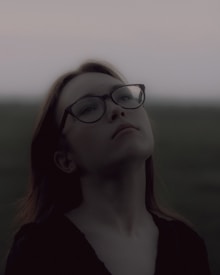 A person with glasses gazes upward with a contemplative expression. The background is softly blurred, suggesting an outdoor setting during twilight or early evening, with subdued lighting.