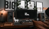 A stylish workspace featuring a large monitor displaying the phrase 'DO MORE' in the center. Surrounding the monitor are various decorative elements, including framed wall art and green ivy. Large speakers flank the monitor on both sides, and there is a classic, warmly lit light bulb on the left. A keyboard, a car model, and a few books are neatly arranged on the wooden desk.