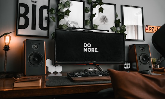 A stylish workspace featuring a large monitor displaying the phrase 'DO MORE' in the center. Surrounding the monitor are various decorative elements, including framed wall art and green ivy. Large speakers flank the monitor on both sides, and there is a classic, warmly lit light bulb on the left. A keyboard, a car model, and a few books are neatly arranged on the wooden desk.