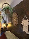 A cozy room features a wicker basket hanging plant, a tapestry with intricate patterns on the wall, and a round mirror with a sunburst frame. The lighting is warm, with a textured lamp casting soft light. A macrame wall hanging adds a bohemian touch, and there are several green plants enhancing the natural feel.
