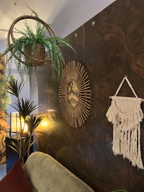 A cozy room features a wicker basket hanging plant, a tapestry with intricate patterns on the wall, and a round mirror with a sunburst frame. The lighting is warm, with a textured lamp casting soft light. A macrame wall hanging adds a bohemian touch, and there are several green plants enhancing the natural feel.