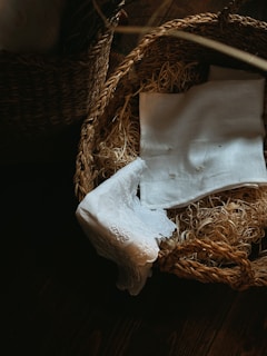 A woven basket is filled with straw and covered with neatly folded white linens. The basket is placed on a dark wooden surface, and the lighting creates a warm, cozy ambiance.