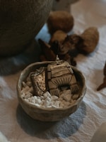 A small ceramic bowl filled with white pebbles holds a decorative, abstract wooden or ceramic piece resembling clothing. It's set on a textured fabric surface, surrounded by rustic, natural materials.