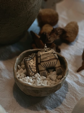 A small ceramic bowl filled with white pebbles holds a decorative, abstract wooden or ceramic piece resembling clothing. It's set on a textured fabric surface, surrounded by rustic, natural materials.