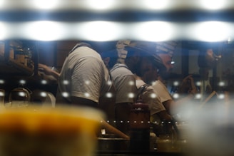 A group of people working in a dimly lit kitchen environment, with reflective surfaces creating a moody and atmospheric ambiance. They are focused on tasks like cooking or preparing food, with warm lights adding a cozy feeling.