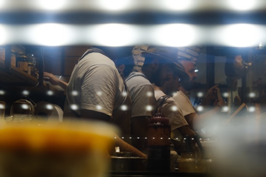 A group of people working in a dimly lit kitchen environment, with reflective surfaces creating a moody and atmospheric ambiance. They are focused on tasks like cooking or preparing food, with warm lights adding a cozy feeling.