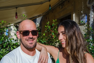 A man wearing sunglasses and a woman with long brown hair are smiling, sitting closely together. They are outdoors with green plants in the background, and the sunlight casts a warm glow on them. The woman rests her arm on the man's shoulder, indicating a sense of closeness.