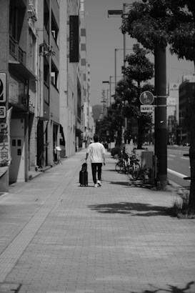 A person wearing a white shirt and black pants is walking down a sidewalk while pulling a suitcase. The street is lined with buildings, trees, and bicycles parked near a signpost. The scene is captured in black and white, giving it a nostalgic or timeless feel.