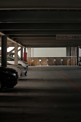 A dimly lit parking garage with several parked cars on the left side under a low ceiling supported by pillars. The structure seems enclosed, with a wall at the back containing some grills and barriers. Overhead signage in a foreign language is visible.