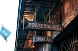 A vintage-looking sign hangs from the ceiling, labeled 'Preservation Hall.' The sign is made of metal, with an aged patina suggesting history and tradition. A blue flag with a white design can be seen flying in the background against a wooden structure with ornate scrollwork.