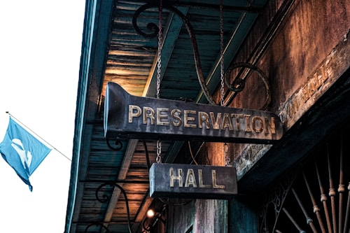 A vintage-looking sign hangs from the ceiling, labeled 'Preservation Hall.' The sign is made of metal, with an aged patina suggesting history and tradition. A blue flag with a white design can be seen flying in the background against a wooden structure with ornate scrollwork.