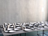 A table is covered with an arrangement of white ceramic dishes and cups of various shapes and sizes, set against a light gray textured wall.