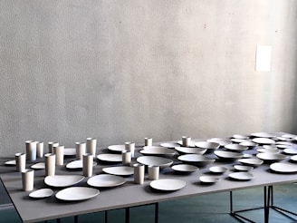 A table is covered with an arrangement of white ceramic dishes and cups of various shapes and sizes, set against a light gray textured wall.