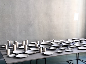 A table is covered with an arrangement of white ceramic dishes and cups of various shapes and sizes, set against a light gray textured wall.