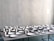 A table is covered with an arrangement of white ceramic dishes and cups of various shapes and sizes, set against a light gray textured wall.