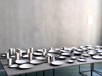 A table is covered with an arrangement of white ceramic dishes and cups of various shapes and sizes, set against a light gray textured wall.