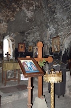 An interior of an ancient stone-walled chapel features religious iconography. There are wooden crosses, framed icons depicting saints, and a golden candle stand holding several lit candles. Dim lighting adds to the historic and sacred atmosphere.