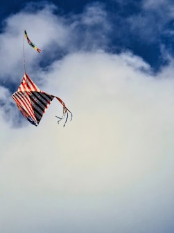 A colorful kite with a pattern of red, white, and blue stripes and trailing ribbons flying against a backdrop of blue sky and clouds.