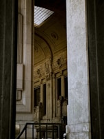Elegant architectural details with intricate carvings and large windows are partially visible, framed by tall stone pillars. A skylight lets in natural light, highlighting the craftsmanship of the interior space.