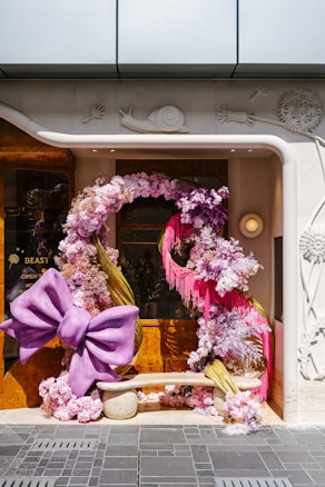 A whimsical storefront display featuring a large purple bow and an abundance of pink and white flowers arranged in an artistic fashion. The flowers form an arch around a beige bench. The decor is set against a polished wooden and stone background with intricate carvings, including a snail motif.
