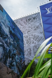 Large wall murals depict abstract art and a detailed map design, with prominently displayed brand logos. The foreground includes green palm leaves, while the sky is overcast.