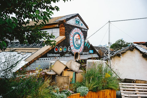 A rustic building with colorful graffiti, featuring a large circular design with geometric patterns and words like 'darkroom' and 'wreck'. The roof is steep and adorned with artistic elements. There are plants and greenery alongside the structure, adding a natural touch amid scattered objects and wooden pallets.
