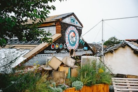 A rustic building with colorful graffiti, featuring a large circular design with geometric patterns and words like 'darkroom' and 'wreck'. The roof is steep and adorned with artistic elements. There are plants and greenery alongside the structure, adding a natural touch amid scattered objects and wooden pallets.