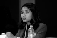 A young person with medium-length hair appears focused while sitting at a table. They are dressed in formal attire, possibly a blazer. In front of them is a partially visible plastic bottle, which might be water. The background is dark, creating a contrast with their lighter clothing.