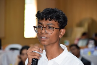 A young person with short curly hair and large glasses is holding a microphone, appearing to speak or present. The person is wearing a white shirt and is situated in a blurred indoor environment, possibly a conference or seminar.