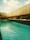 A serene swimming pool surrounded by a wooden fence. The clear blue water reflects the partly cloudy sky above. Small potted plants and a bamboo screen add a touch of natural beauty to the scene.