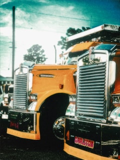 A vibrant, stylized image of a large, shiny yellow truck with chrome details parked outdoors. The truck features a prominent grille and headlights, with a branded logo visible. The background includes a partly cloudy sky and silhouetted trees, adding to the dramatic effect.