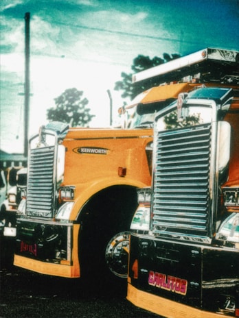 A vibrant, stylized image of a large, shiny yellow truck with chrome details parked outdoors. The truck features a prominent grille and headlights, with a branded logo visible. The background includes a partly cloudy sky and silhouetted trees, adding to the dramatic effect.