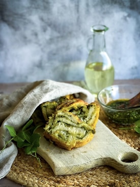A rustic wooden cutting board holds several pieces of bread swirled with a green, possibly pesto, filling. The bread is partially covered with a light beige cloth. Next to the cutting board is a clear glass bowl filled with a green sauce and a wooden spoon. In the background, there is a glass bottle with a handle containing a light yellow liquid, placed on a wooden surface. The background is neutral with soft, slightly textured walls.