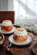 Two plates with cream-filled pastries on a rustic wooden table. The soft, fluffy cream sits atop brown, glossy buns. In the foreground, there are some nuts and small white flowers scattered across the table. A blurred, light-filled window provides a gentle background.