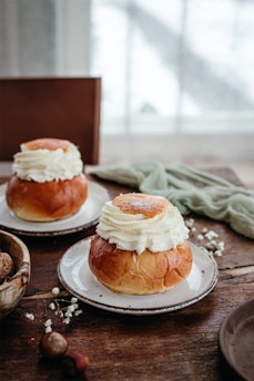 Two plates with cream-filled pastries on a rustic wooden table. The soft, fluffy cream sits atop brown, glossy buns. In the foreground, there are some nuts and small white flowers scattered across the table. A blurred, light-filled window provides a gentle background.