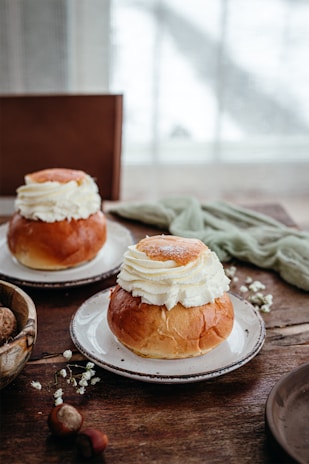 Two plates with cream-filled pastries on a rustic wooden table. The soft, fluffy cream sits atop brown, glossy buns. In the foreground, there are some nuts and small white flowers scattered across the table. A blurred, light-filled window provides a gentle background.
