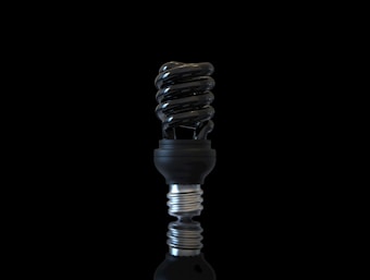 A modern, spiral-shaped light bulb is depicted against a black background. The bulb has a sleek, metallic finish with a reflective surface that catches light, emphasizing its clean and futuristic design. The compact fluorescent lamp is positioned vertically, showcasing its distinct coiled structure.