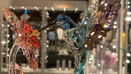 A close-up view of a beautifully crafted crystal sculpture depicting a hummingbird feeding from a flower. The hummingbird is made with a range of colors including blue, green, and brown. The flower has vibrant red, yellow, and pink hues. The background appears blurry with sparkles, likely reflecting off other crystal items displayed behind glass.