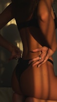 A person stands with their back turned, wearing dark underwear. The lighting creates dramatic shadows, accentuating the body's curves and contours, with a hand seen in the frame gesturing in a relaxed manner.