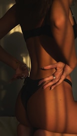 A person stands with their back turned, wearing dark underwear. The lighting creates dramatic shadows, accentuating the body's curves and contours, with a hand seen in the frame gesturing in a relaxed manner.