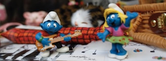 Two small, blue, animated figures with white hats are placed on a surface filled with assorted items. One is playing a guitar, while the other is raising a hand in a playful gesture. Behind them are objects including a plush toy and wicker basket, adding a whimsical atmosphere.