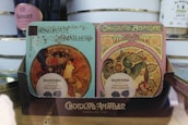 Two ornately designed chocolate boxes with Art Nouveau style illustrations featuring women adorned with floral and decorative elements. The packaging indicates 'Chocolate Amatller' from Barcelona with 70% cacao content. The design is intricate and vintage in appearance, placed on a wooden surface with other items visible in the background.