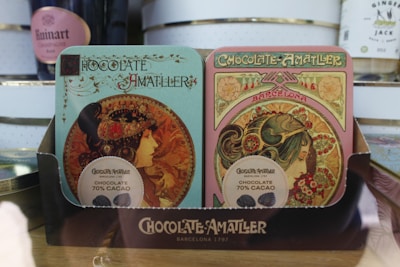 Two ornately designed chocolate boxes with Art Nouveau style illustrations featuring women adorned with floral and decorative elements. The packaging indicates 'Chocolate Amatller' from Barcelona with 70% cacao content. The design is intricate and vintage in appearance, placed on a wooden surface with other items visible in the background.