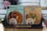 Two ornately designed chocolate boxes with Art Nouveau style illustrations featuring women adorned with floral and decorative elements. The packaging indicates 'Chocolate Amatller' from Barcelona with 70% cacao content. The design is intricate and vintage in appearance, placed on a wooden surface with other items visible in the background.