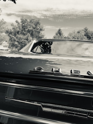 A black vintage car's rear end with a prominent 'Chevrolet' emblem. The car is positioned against a backdrop of trees and a partly cloudy sky. The image is captured in black and white, giving it a nostalgic feel.