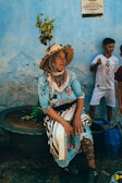 An elderly woman is sitting beside a large planter with a small tree. She wears a colorful floral dress and a decorative straw hat. A child stands nearby, wearing a white sports jersey. The wall behind them is painted a vibrant blue with some text and flags visible on a poster.