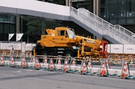 A large yellow crane vehicle is parked in an urban construction area. Traffic cones and barriers are arranged in front of it, and a white pedestrian bridge is visible above. Surrounding buildings add to the metropolitan setting.