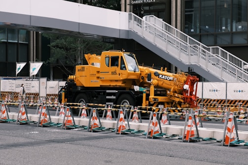 A large yellow crane vehicle is parked in an urban construction area. Traffic cones and barriers are arranged in front of it, and a white pedestrian bridge is visible above. Surrounding buildings add to the metropolitan setting.