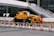 A large yellow crane vehicle is parked in an urban construction area. Traffic cones and barriers are arranged in front of it, and a white pedestrian bridge is visible above. Surrounding buildings add to the metropolitan setting.